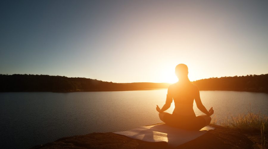 Silhouette of healthy woman is practicing yoga at mountain lake during sunset.